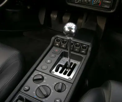 Interior of a black car showing a chrome manual gear shifter, center console with buttons and climate controls.