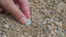 Close-up of a hand picking up a small silver coin from gravelly ground outdoors.