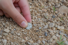 Close-up of a hand picking up a small silver coin from gravelly ground outdoors.
