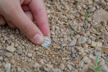Close-up of a hand picking up a small silver coin from gravelly ground outdoors.
