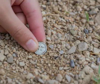 Close-up of a hand picking up a small silver coin from gravelly ground outdoors.