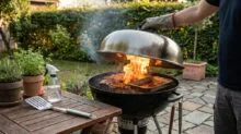 Person lifting a blazing kettle grill lid to charred meat on the grate outdoors, with a wooden table and potted herbs nearby.