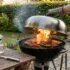 Person lifting a blazing kettle grill lid to charred meat on the grate outdoors, with a wooden table and potted herbs nearby.