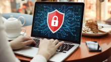 Person typing on a laptop displaying a red shield with a white lock over a digital circuit background, in a cafe with a smartphone nearby.