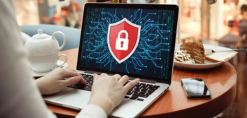 Person typing on a laptop displaying a red shield with a white lock over a digital circuit background, in a cafe with a smartphone nearby.