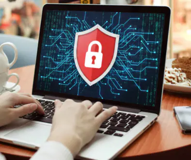 Person typing on a laptop displaying a red shield with a white lock over a digital circuit background, in a cafe with a smartphone nearby.