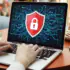 Person typing on a laptop displaying a red shield with a white lock over a digital circuit background, in a cafe with a smartphone nearby.