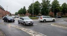 Silver Renault hatchback in the foreground navigating a wet roundabout, with other cars and brick row houses with trees in the background.