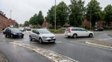 Silver Renault hatchback in the foreground navigating a wet roundabout, with other cars and brick row houses with trees in the background.