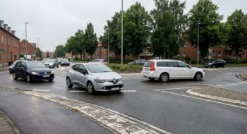 Silver Renault hatchback in the foreground navigating a wet roundabout, with other cars and brick row houses with trees in the background.
