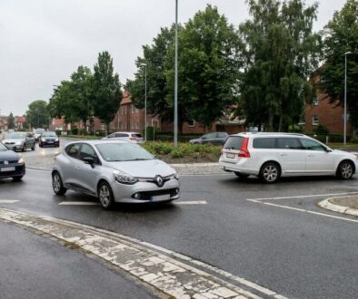 Silver Renault hatchback in the foreground navigating a wet roundabout, with other cars and brick row houses with trees in the background.