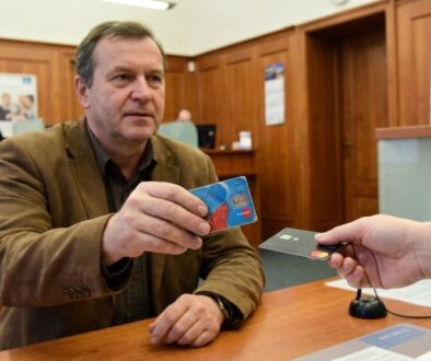 Man in a brown jacket at a bank counter hands a blue debit card toward the clerk during a transaction.