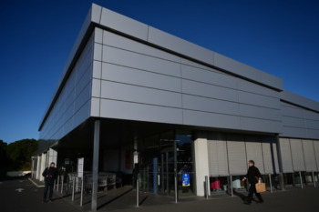 Modern gray commercial building with a glass entrance, set against a deep blue sky, two people outside—one with a cart, another carrying a shopping bag.