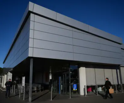 Modern gray commercial building with a glass entrance, set against a deep blue sky, two people outside—one with a cart, another carrying a shopping bag.
