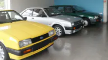 Row of vintage Ford cars in a showroom: yellow car in front, followed by silver and green cars lined up against a blue wall.