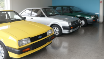 Row of vintage Ford cars in a showroom: yellow car in front, followed by silver and green cars lined up against a blue wall.