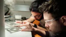 Two researchers in safety glasses lean over a lab bench, examining a glass slide or small sample under bright light.