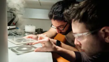 Two researchers in safety glasses lean over a lab bench, examining a glass slide or small sample under bright light.