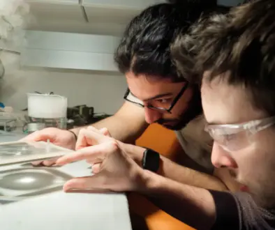 Two researchers in safety glasses lean over a lab bench, examining a glass slide or small sample under bright light.