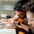 Two researchers in safety glasses lean over a lab bench, examining a glass slide or small sample under bright light.