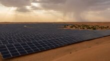 Rows of dark solar panels stretch across a desert landscape under a cloudy sky with sunbeams breaking through.