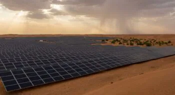 Rows of dark solar panels stretch across a desert landscape under a cloudy sky with sunbeams breaking through.