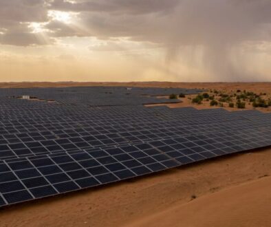 Rows of dark solar panels stretch across a desert landscape under a cloudy sky with sunbeams breaking through.