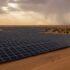 Rows of dark solar panels stretch across a desert landscape under a cloudy sky with sunbeams breaking through.