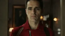 Man in a red jacket with suspenders stands in a doorway, looking straight ahead with a serious expression.