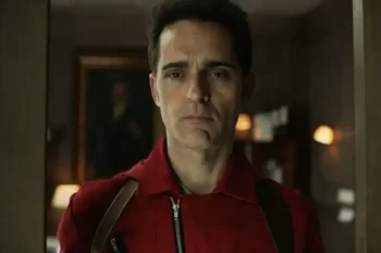 Man in a red jacket with suspenders stands in a doorway, looking straight ahead with a serious expression.