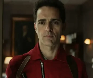 Man in a red jacket with suspenders stands in a doorway, looking straight ahead with a serious expression.