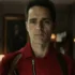 Man in a red jacket with suspenders stands in a doorway, looking straight ahead with a serious expression.