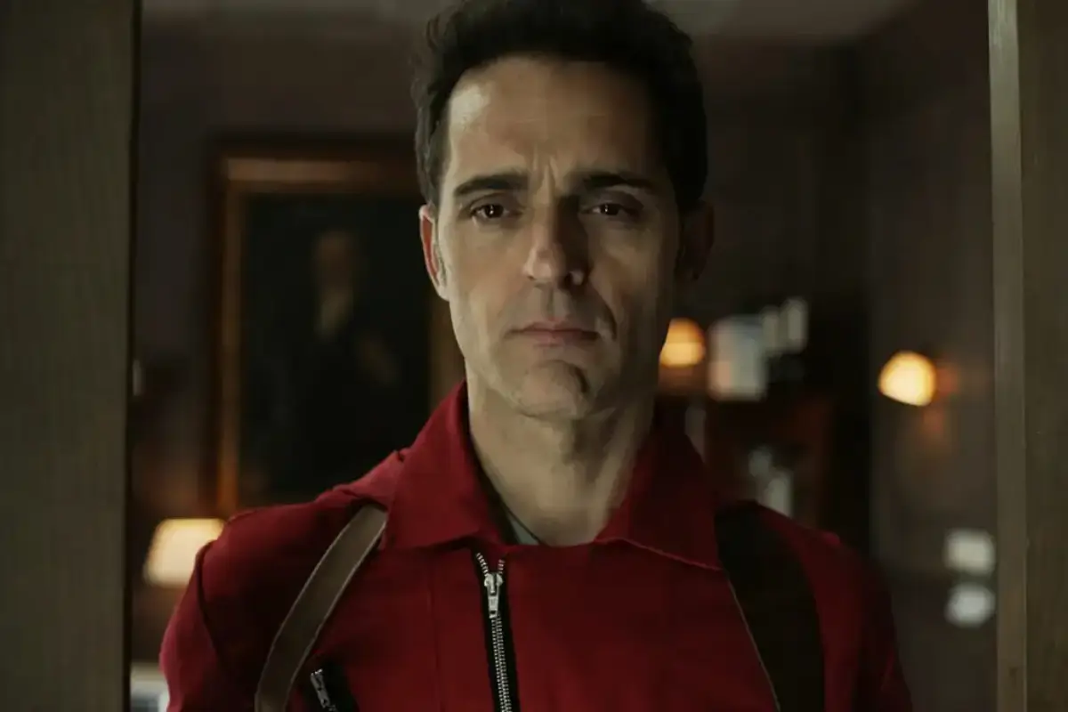 Man in a red jacket with suspenders stands in a doorway, looking straight ahead with a serious expression.