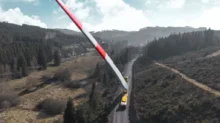 A giant red-and-white wind turbine blade spans a rural road, supported by trucks amid hills and forests.