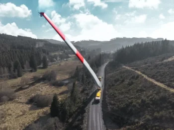 A giant red-and-white wind turbine blade spans a rural road, supported by trucks amid hills and forests.