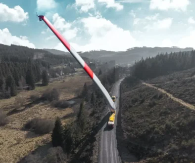 A giant red-and-white wind turbine blade spans a rural road, supported by trucks amid hills and forests.