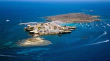 A rocky island town with white houses around a small harbor, boats anchored in blue sea.