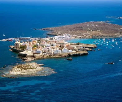 A rocky island town with white houses around a small harbor, boats anchored in blue sea.
