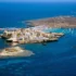 A rocky island town with white houses around a small harbor, boats anchored in blue sea.