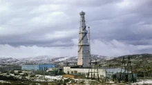 Tall tower with guy wires rising above a cluster of low industrial buildings in a snow-covered, desolate landscape under a cloudy sky.