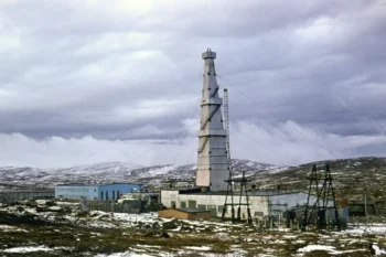 Tall tower with guy wires rising above a cluster of low industrial buildings in a snow-covered, desolate landscape under a cloudy sky.
