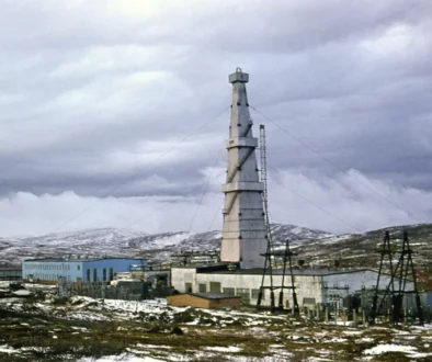 Tall tower with guy wires rising above a cluster of low industrial buildings in a snow-covered, desolate landscape under a cloudy sky.