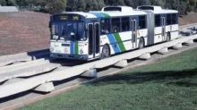Articulated city bus travelling on an elevated concrete guideway beside a grassy area, with trees in the background.