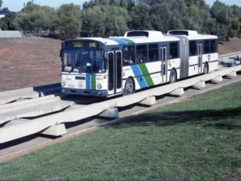 Articulated city bus travelling on an elevated concrete guideway beside a grassy area, with trees in the background.