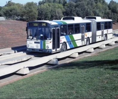Articulated city bus travelling on an elevated concrete guideway beside a grassy area, with trees in the background.