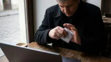 Man in a dark coat at a café table, holding a credit card next to a laptop and a coffee cup on the table.