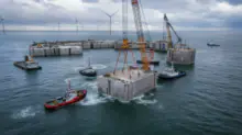 Offshore construction scene with large concrete blocks, cranes, and tugboats in calm sea; wind turbines offshore in the distance