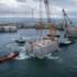 Offshore construction scene with large concrete blocks, cranes, and tugboats in calm sea; wind turbines offshore in the distance