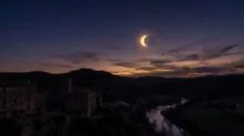 Twilight over a stone village and winding river valley as a solar eclipse forms a bright crescent in the sky behind distant hills.