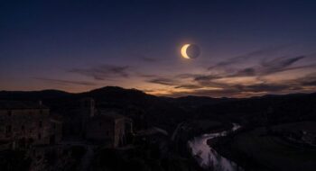 Twilight over a stone village and winding river valley as a solar eclipse forms a bright crescent in the sky behind distant hills.